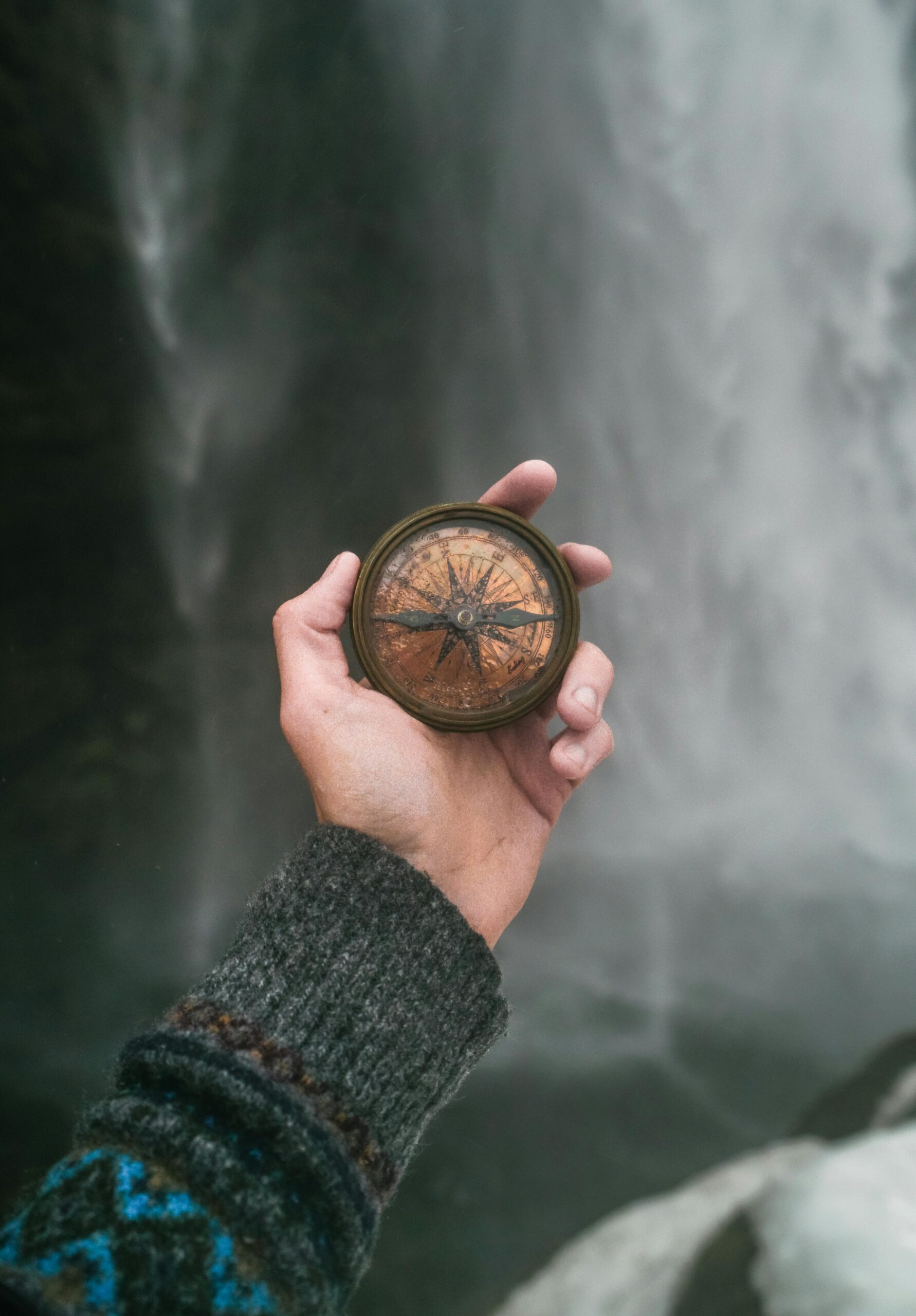 Close-up of a person's hand holding a compass near a waterfall, symbolizing exploration and adventure.