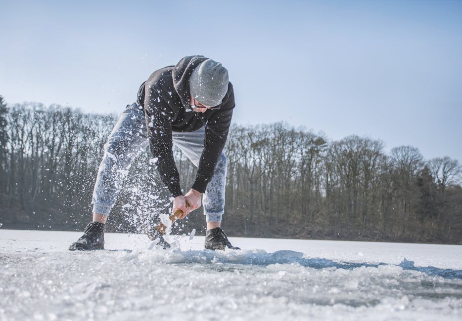 A man chopping ice on a frozen lake surrounded by trees during a cold winter day.